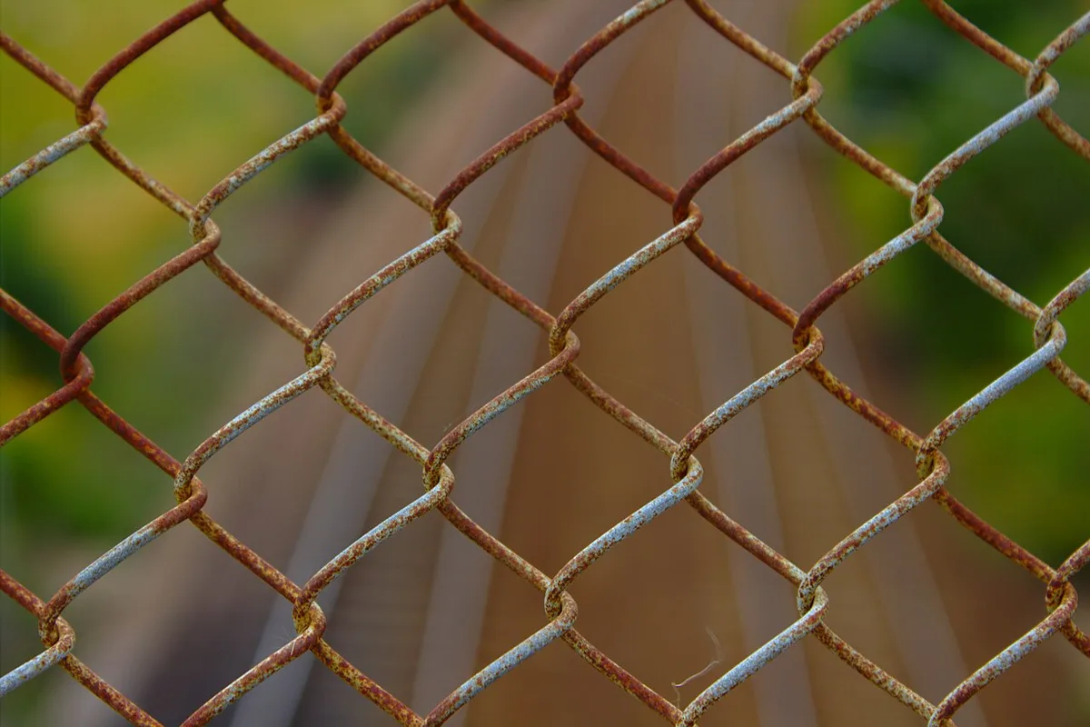 A rusted chain-link fence with railway tracks blurred behind it